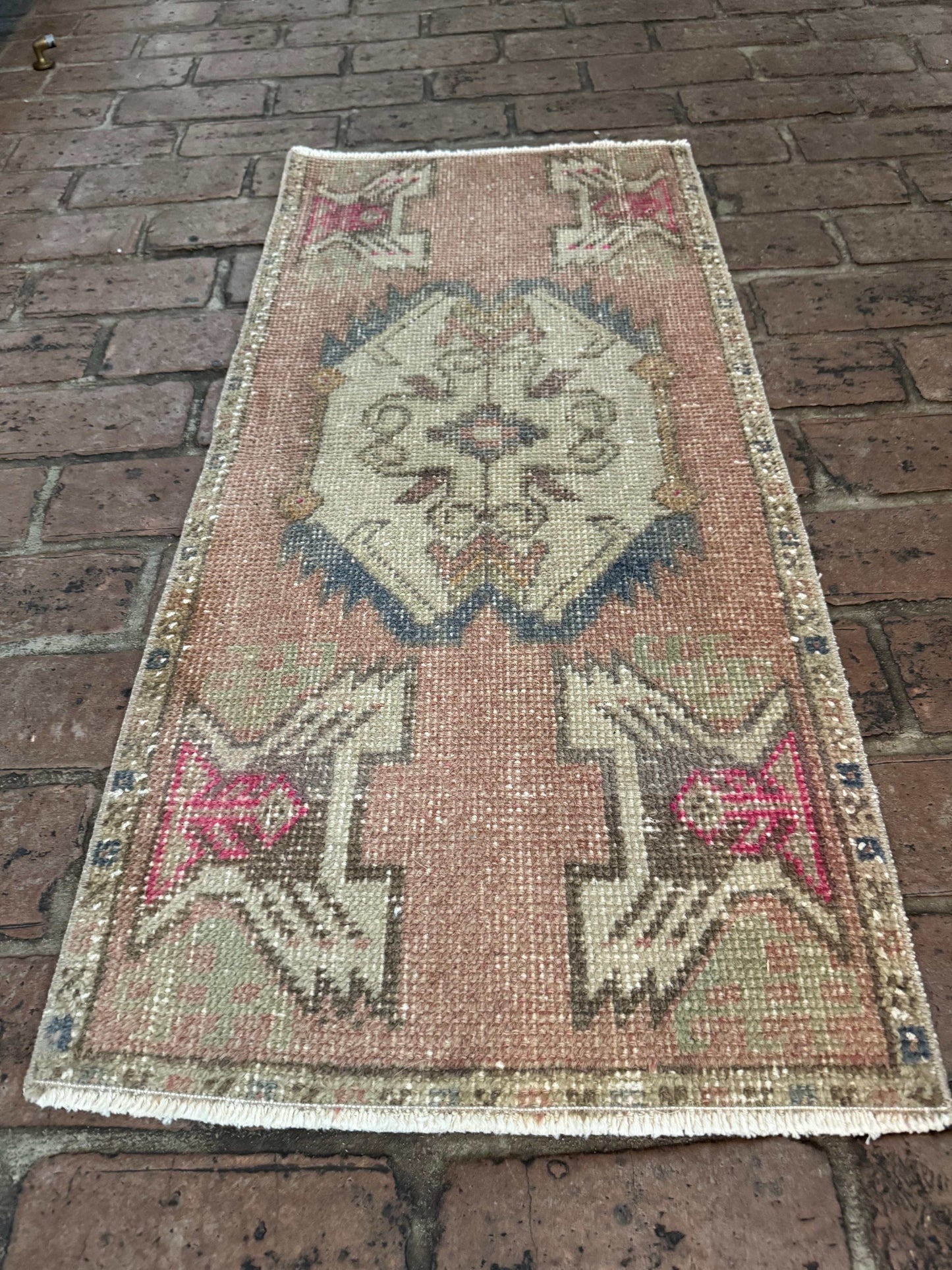 Small Turkish rug mat in mauve pink, gray-blue, brown, and hot pink, measuring 1'6" x 3'3" on brick floor.