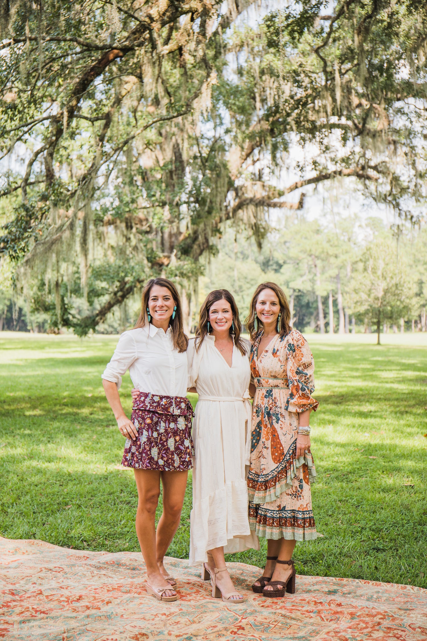 Three women standing outdoors in a park-like setting with trees and green grass.